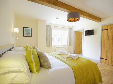 A bedroom with a bed with green cushions and blanket a window wall lamp wooden doors mounted television and exposed wooden beam at Park Stables Wyck Beacon near Bourton-on-the-Water