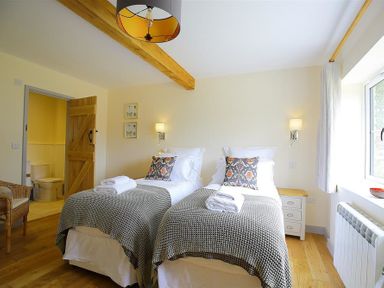 A bedroom with two single beds a wooden door open to a bathroom and a window with curtains at Park Stables in Wyck Beacon near Bourton-on-the-Water
