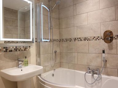 A bathroom with a pedestal sink a lighted mirror a bathtub with a showerhead and tiled walls at Park Stables Wyck Beacon near Bourton-on-the-Water