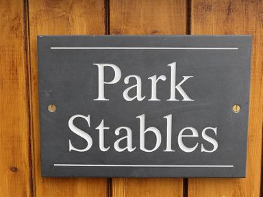 A dark rectangular sign with white text saying Park Stables mounted on a wooden wall at Park Stables Wyck Beacon near Bourton-on-the-Water