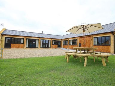 A courtyard with a wooden picnic table and umbrella in front of a single-story building with stone and wood walls at Park Stables Wyck Beacon near Bourton-on-the-Water