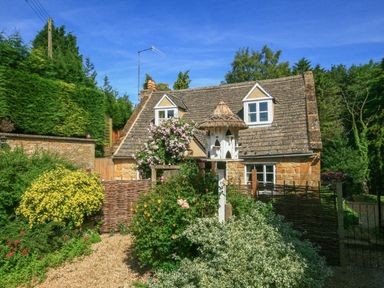 A stone cottage with two dormer windows surrounded by a garden with flowers and a birdhouse at Hadcroft Cottage in Aston Magna