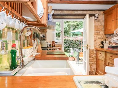A kitchen with wooden cabinets a sink dishes hanging above and an open door to a garden at Hadcroft Cottage in Aston Magna