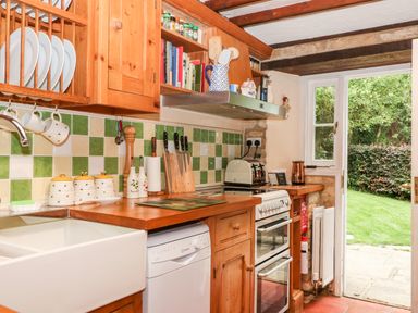A kitchen with wooden cabinets a white sink, dishwasher, stove, toaster, and an open door leading to a garden at Hadcroft Cottage in Aston Magna