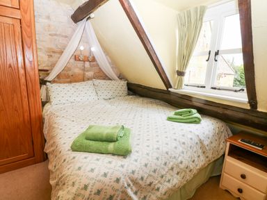 A bedroom with a double bed green towels wooden wardrobe window with curtains and bedside table at Hadcroft Cottage in Aston Magna