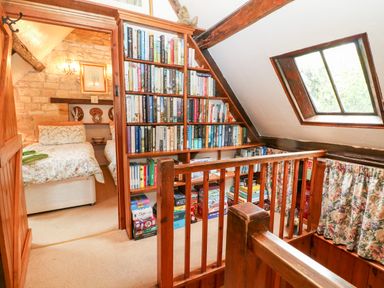 A small landing area with a wooden bookshelf filled with books and board games next to a staircase and a slanted window at Hadcroft Cottage in Aston Magna