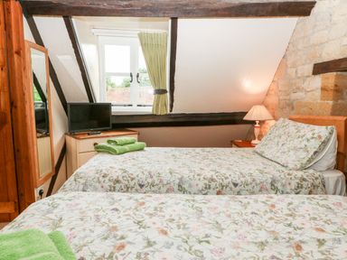 A bedroom with two single beds a bedside table with lamp a TV on a dresser and a window with green curtains at Hadcroft Cottage in Aston Magna