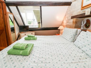 A bedroom with a bed with patterned sheets and green towels a dresser with a television a lamp and a window with green curtains at Hadcroft Cottage in Aston Magna