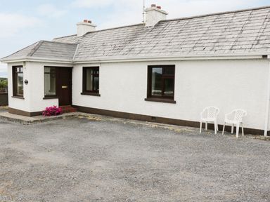 Exterior of a white house with three windows a brown door two white chairs and pink flowers at Daffodil Cottage in Grange County Sligo