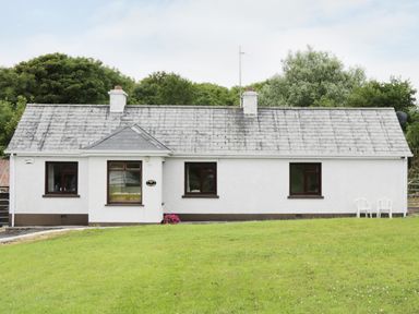 A white single-story house with three windows two chimneys and two white chairs on green grass at Daffodil Cottage in Grange County Sligo