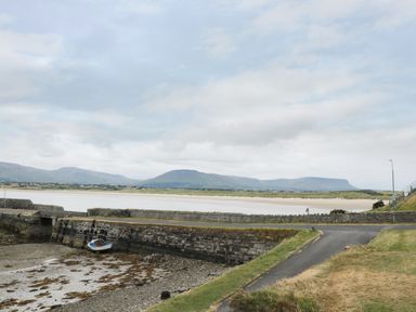 A small boat docked near a stone seawall with mountains and a sandy shore in the background at Daffodil Cottage in Grange County Sligo