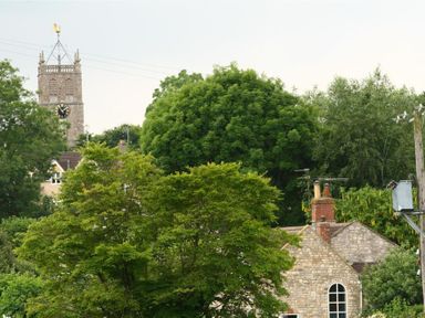 A view of a stone building with chimneys and arched windows behind green trees and a clock tower in the background at Cider Press Cottage Priston Village Bath