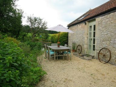 An outdoor patio with a wooden table and chairs under a striped umbrella next to a stone building with plants Cider Press Cottage Priston Village Bath