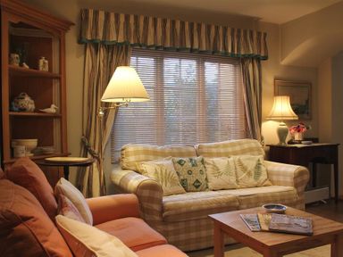 A living room with two sofas a wooden coffee table with magazines a floor lamp a side table a wooden cabinet and a window with blinds and curtains at The Mews Cottage in Tisbury