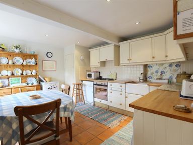 A kitchen with a wooden dining table covered with a checkered tablecloth and wooden chairs a cabinet with dishes a counter with a stove microwave and sink and striped rugs on the floor at The Mews Cottage in Tisbury