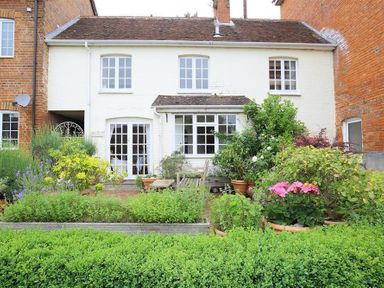 A garden with various plants and flowers in front of a white two-story house with multiple windows and a wooden bench at The Mews Cottage in Tisbury