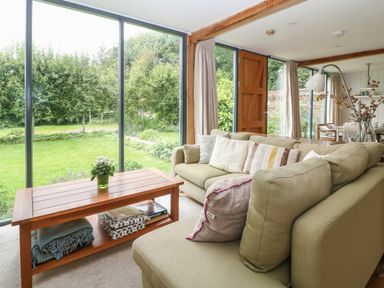 A living room with a green sofa and wooden coffee table by large glass windows showing a garden at The Cart Barn in Ridge near Chilmark