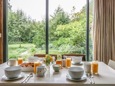 A dining table set with bowls mugs glasses of orange juice and pastries in front of a large window showing a green garden at The Cart Barn in Ridge near Chilmark