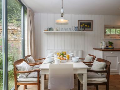 A dining area with a white table set with bowls glasses and croissants four wooden chairs with cushions and a large window at The Cart Barn in Ridge near Chilmark