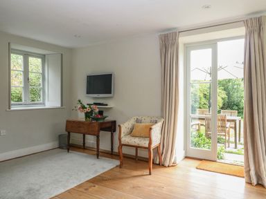 A room with a wooden floor a small wooden table a floral armchair a wall-mounted TV and glass doors leading to a garden at The Cart Barn Ridge near Chilmark