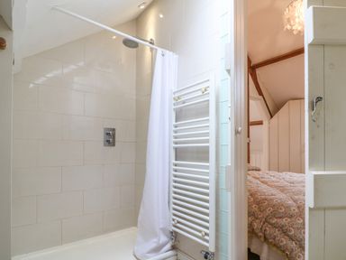 A bathroom with a shower area and white shower curtain next to a heated towel rail and a view into a bedroom with patterned bedding at The Cart Barn in Ridge near Chilmark