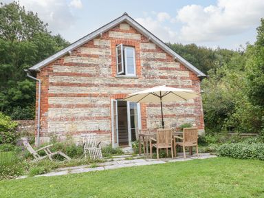 A stone and brick house with an open door and window a patio table with an umbrella and chairs in a garden at The Cart Barn in Ridge near Chilmark