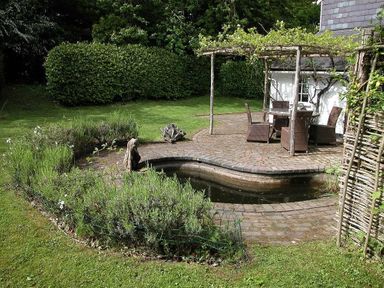 A garden with a small pond a paved patio with a table and four chairs under a wooden pergola at Gun Hill Cottage in Heathfield