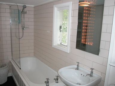 A bathroom with a bathtub and shower, a sink with two taps, a window with blinds and a mirror with lights at Gun Hill Cottage in Heathfield