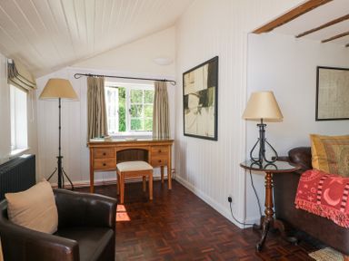 A room with a wooden desk and stool in front of a window a black leather chair a small round table with a lamp and a sofa with cushions and a blanket at Gun Hill Cottage in Heathfield