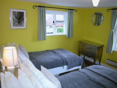 A bedroom with two beds covered in gray blankets a lamp on a bedside table a small chest of drawers a round mirror and windows with light curtains at Gun Hill Cottage in Heathfield