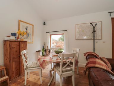 A dining area with a wooden table covered with a checked tablecloth surrounded by four white chairs with cushions a wooden cabinet with a pitcher and flowers a framed picture on the wall a coat rack a brown leather sofa with a blanket and a window showing greenery at Russet in Martin