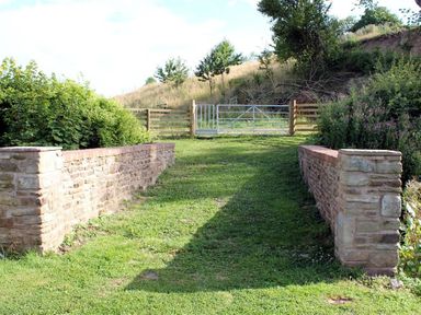 A grassy pathway with stone walls on both sides leading to a metal gate and wooden fencing at The Old Dairy in Flaxley