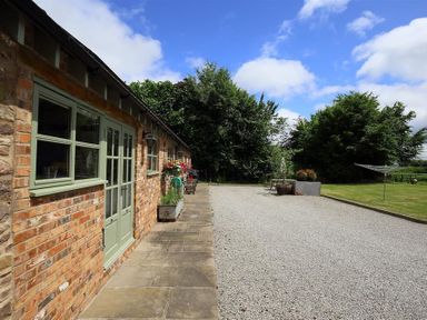 A brick building with green windows and doors next to a gravel driveway and a grassy area with trees at The Old Dairy in Flaxley