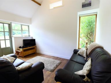 A living room with two black leather sofas a wooden TV stand with a television and windows showing greenery outside at The Old Dairy in Flaxley
