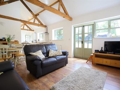 A living room with wooden beams a black leather sofa a wooden dining table and green French doors at The Old Dairy in Flaxley