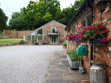 An outdoor area with gravel driveway flower baskets on brick walls and green trees at The Old Dairy in Flaxley