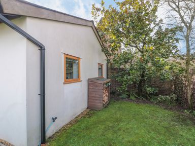 A side garden area with two windows on a white wall a wooden log storage shed and a tree next to a woven fence at Pippin in Martin
