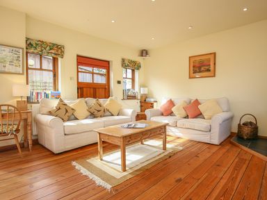 A living room with two sofas, a wooden coffee table, wooden floors, and windows with curtains at The Coach House in Lympstone