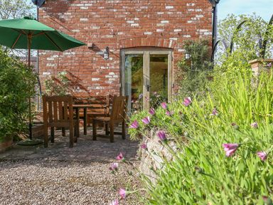 An outdoor patio with wooden chairs and table under a green umbrella next to a brick wall with glass doors and plants with purple flowers at The Coach House in Lympstone