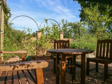 An outdoor patio with wooden chairs and a round table surrounded by greenery and brick walls at The Coach House in Lympstone