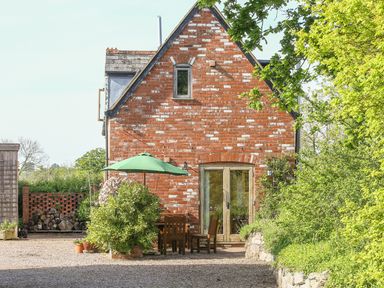 A brick house with a green umbrella and wooden table and chairs outside surrounded by greenery at The Coach House in Lympstone