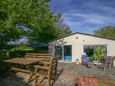 An outdoor patio area with wooden table and benches under a tree and two purple lounge chairs in front of a building with sliding glass doors at The Coach House in Lympstone