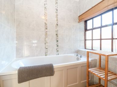 A bathroom with a white bathtub grey tiled walls a wooden shelf unit and a large window with a blind at Spring Cottage in Wroxton