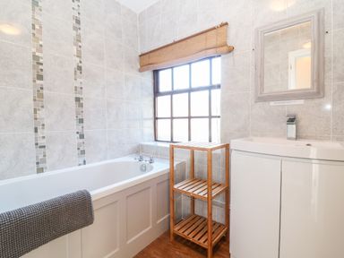 A bathroom with a bathtub a wooden shelf a window with a blind a sink with a cabinet and a mirror at Spring Cottage in Wroxton