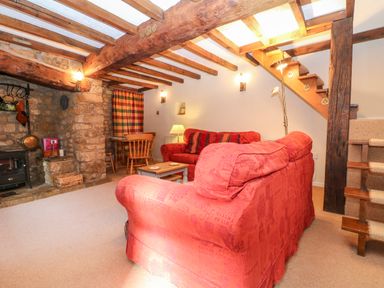 A living room with red sofas a wooden dining table and chairs a stone fireplace and exposed wooden beams at Spring Cottage in Wroxton