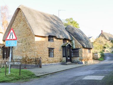 A stone cottage with a thatched roof next to a road with traffic signs and a Mills Lane street sign at Spring Cottage in Wroxton