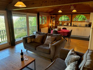 A living room with sofas wine bottle and glasses dining table kitchen area and large windows at Treetops in Woody Bay