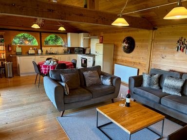 A living area with grey sofas and a wooden coffee table with a wine bottle and glass next to a kitchen with white cabinets and a round table covered with a red checkered tablecloth at Treetops in Woody Bay