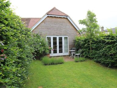 A garden with grass shrubs a small tree a white table and two chairs and a wooden house with glass doors at Bridge Studio in Broad Chalke