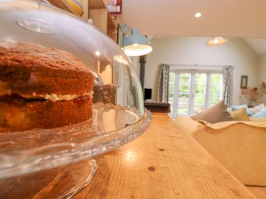 A cake on a glass cake stand on a wooden table in a living room with a sofa and large windows at Bridge Studio in Broad Chalke